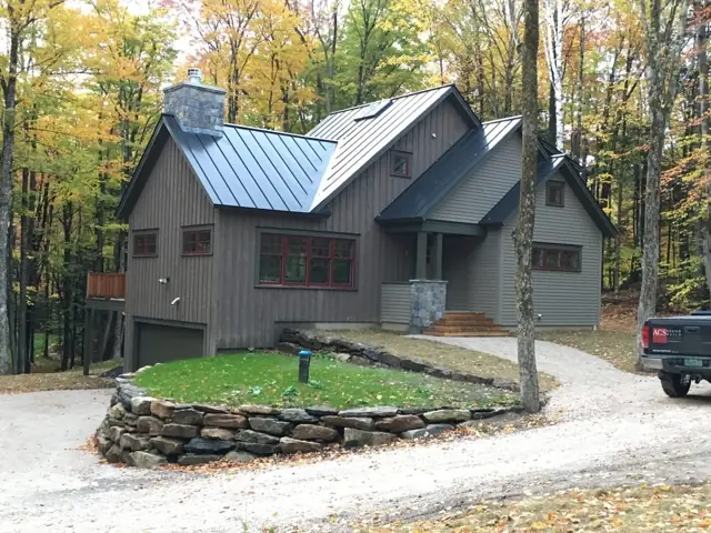 Gray board-and-batten home with metal roof and stone retaining wall in autumn